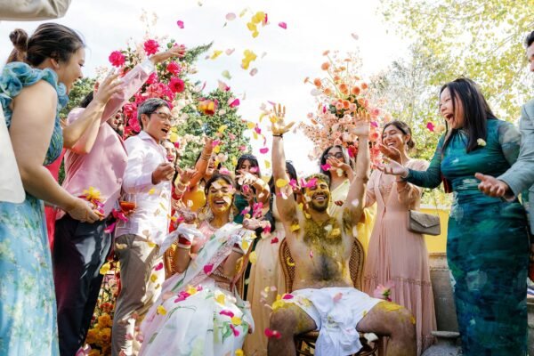 A bride and groom laugh as family and guests shower them with vibrant yellow and pink flower petals during an outdoor Haldi ceremony in Spain.