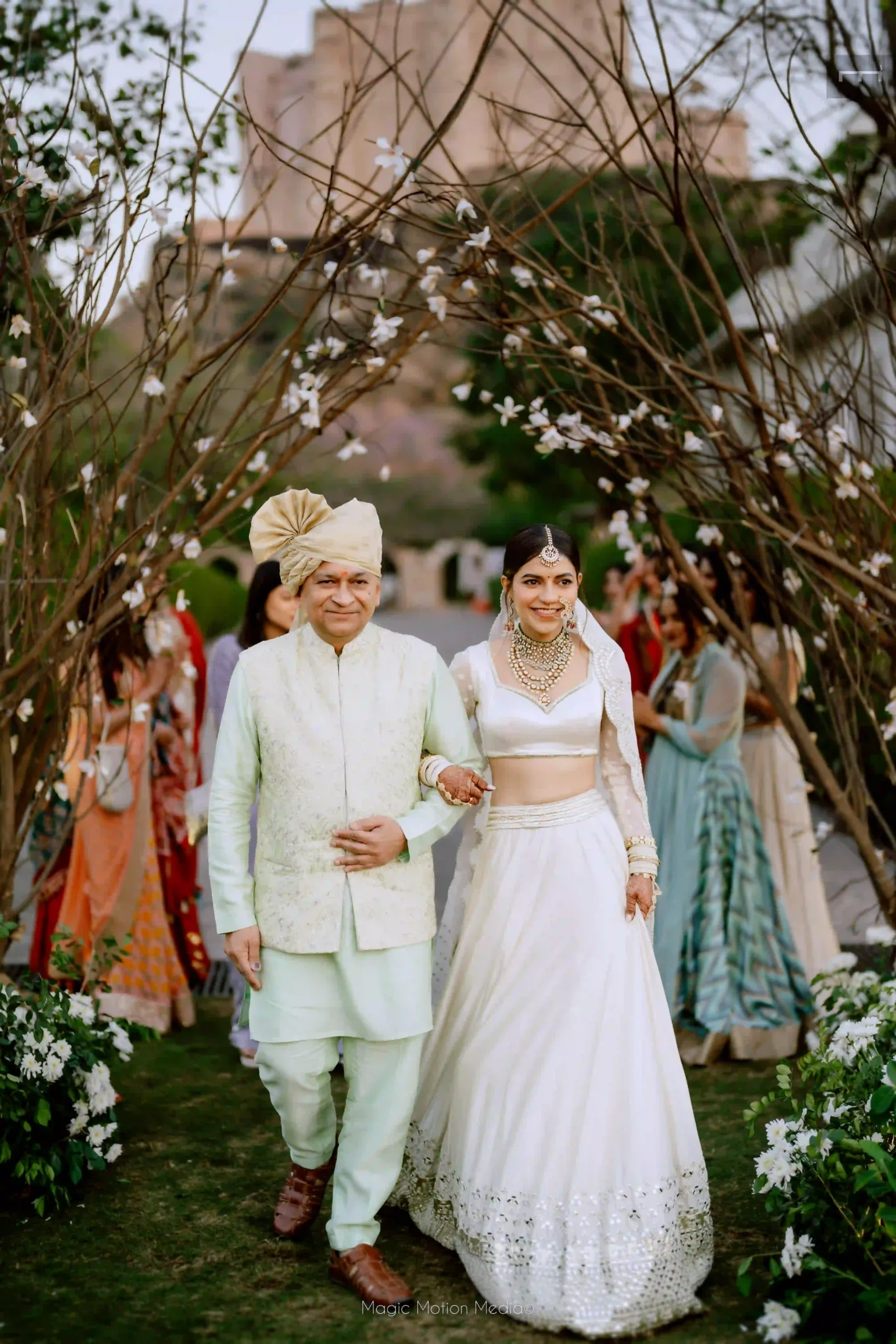 Bride in an ivory lehenga walking arm in arm with an older gentleman beneath flowering branches at Alila Fort Bishangarh.