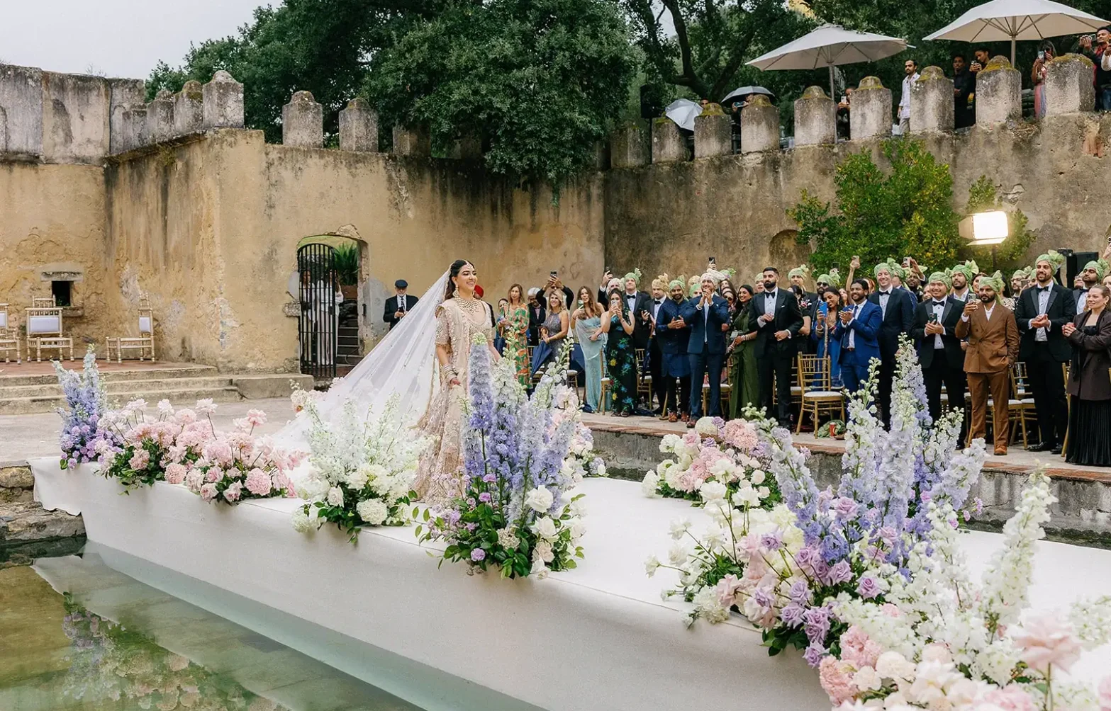 3. An Indian bride in an embellished lehenga walks down a white aisle bordered by pastel purple and pink flowers over a pool at Penha Longa Resort in Sintra, Portugal while guests look on.