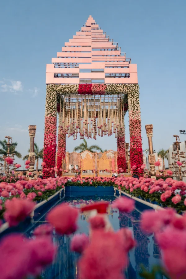 A tall geometric tiered mandap with pillars covered in a red, pink, and white floral ombre gradient stands at the end of a long reflective water aisle bordered by pink roses.