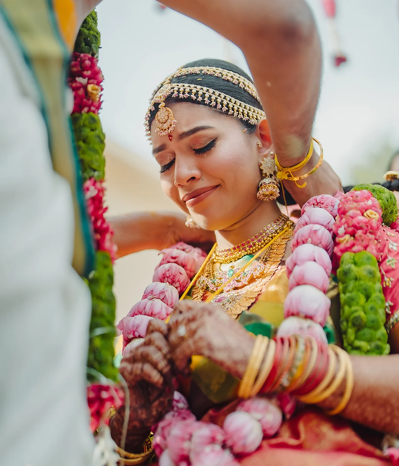 Bride with eyes closed as a ceremonial thread is tied around her neck, wearing traditional gold jewellery and pink-and-green floral garlands.