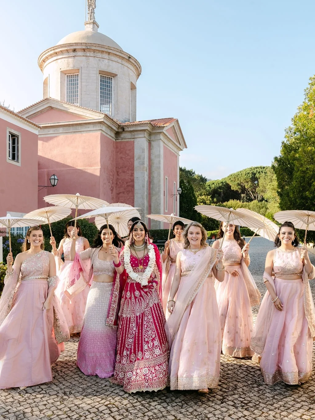 A bride in a magenta lehenga walks with her bridesmaids holding white parasols at the Penha Longa Resort in Sintra, Portugal, featured in the international wedding destinations gallery.