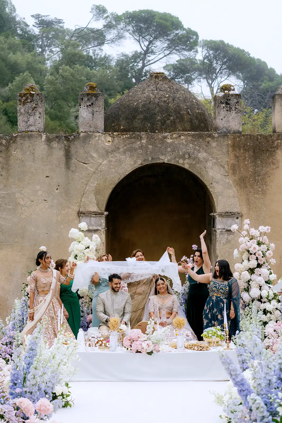 Bride and groom seated beneath a white ceremonial cloth, surrounded by loved ones and pastel flowers at their Indian wedding in Portugal.