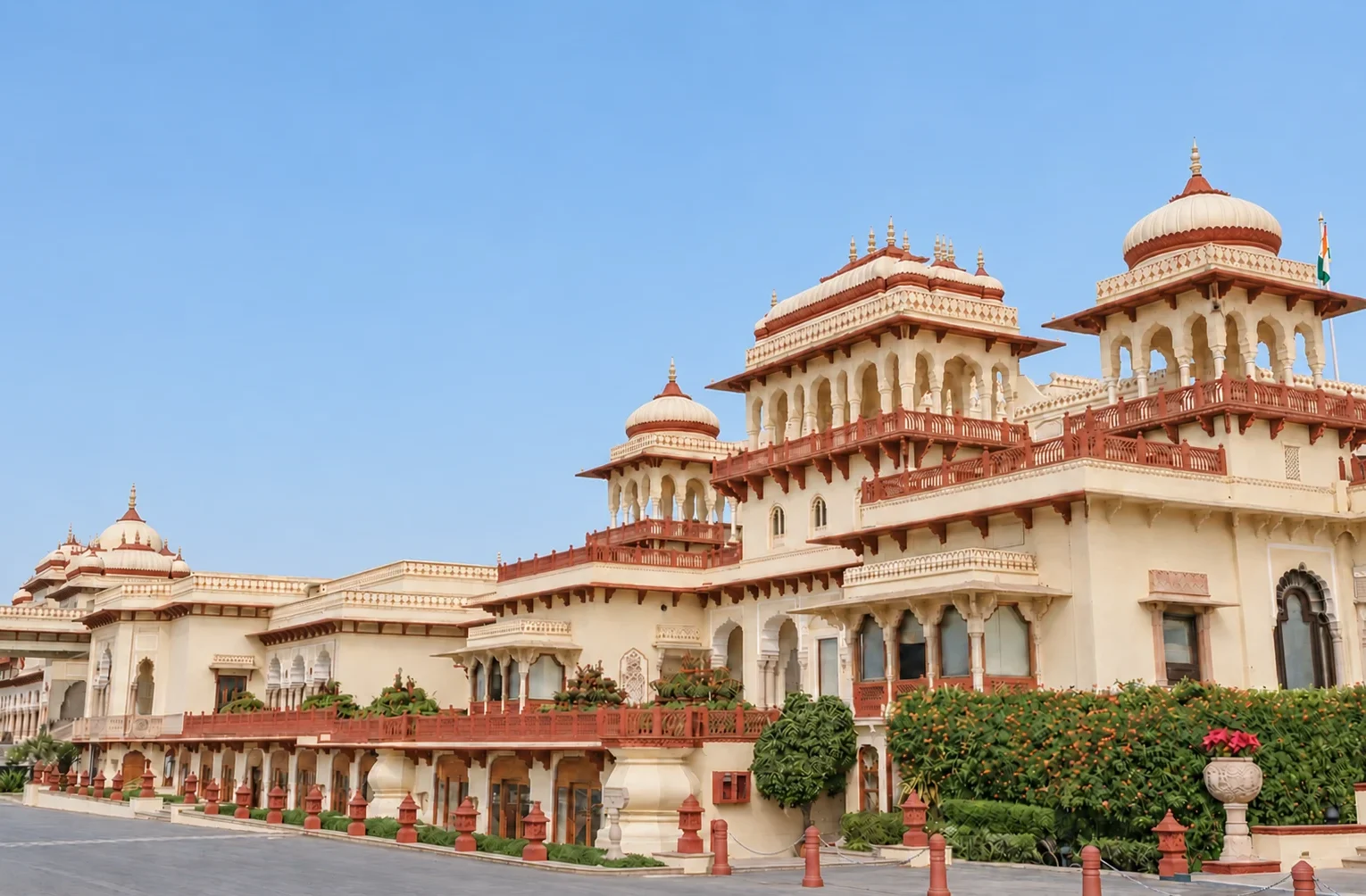 The grand exterior architecture of Rambagh Palace in Jaipur features traditional jharokhas, chhatris, and red sandstone railings under a clear blue sky.