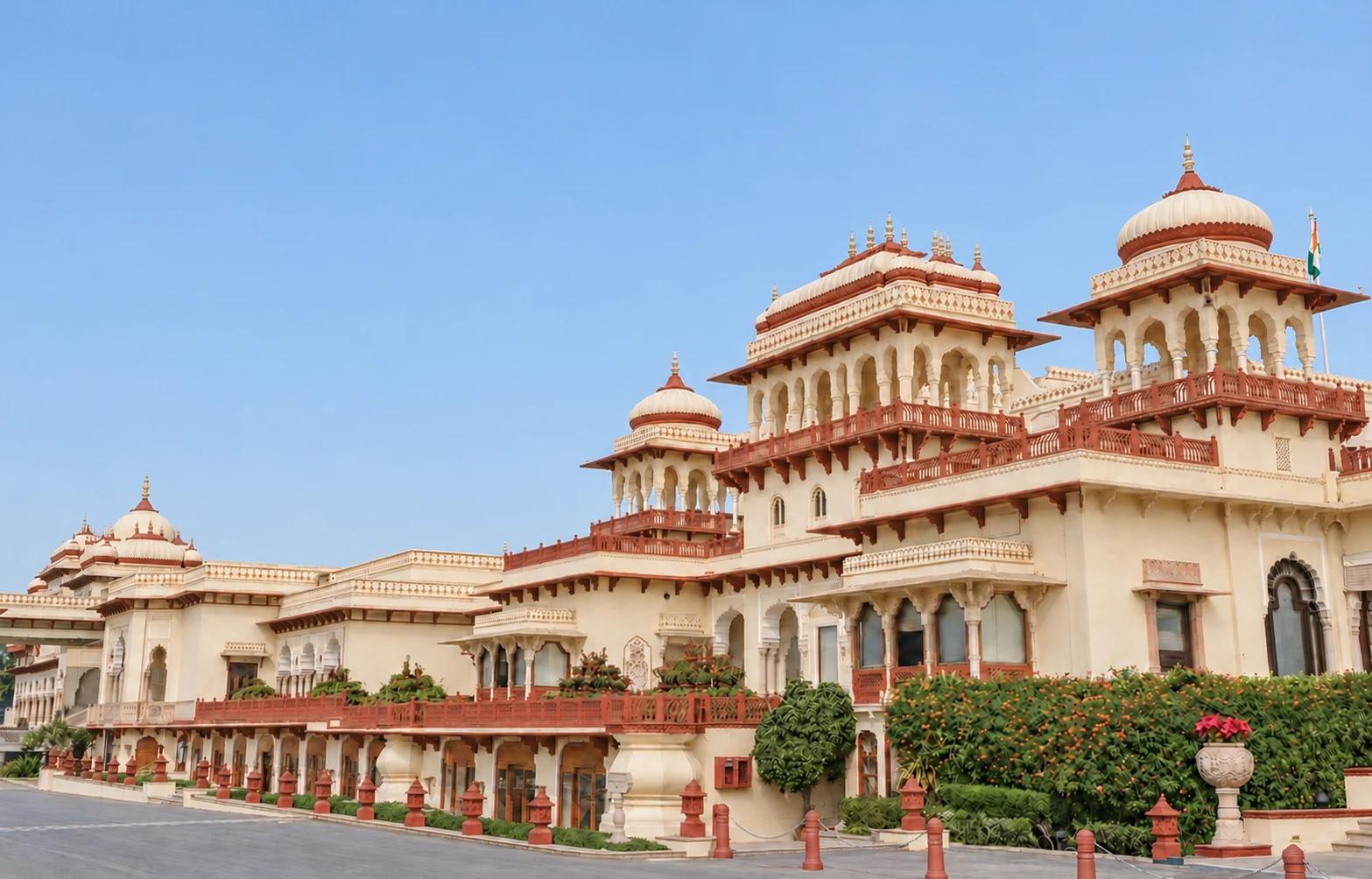 The grand exterior architecture of Rambagh Palace in Jaipur features traditional jharokhas, chhatris, and red sandstone railings under a clear blue sky.