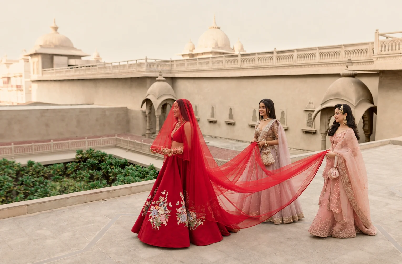 3. A bride in a red Sabyasachi lehenga walks on an outdoor stone terrace at Fairmont Jaipur while two bridesmaids carry her trailing red veil against traditional palatial domes.