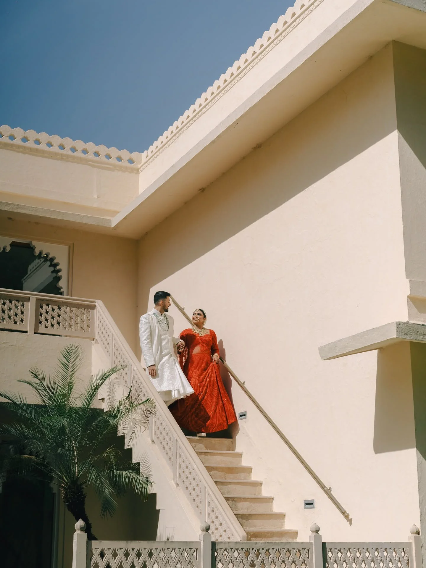 A bride in a bright red lehenga and a groom in a white sherwani walk down an outdoor beige stone staircase beneath a clear blue sky at Trident Hotel Udaipur.