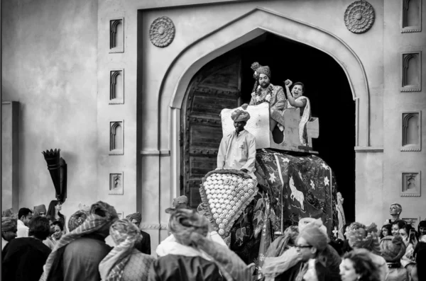A black and white photograph captures a groom and a co-passenger riding a beautifully decorated ceremonial elephant out from beneath a massive traditional archway surrounded by wedding guests in Jaipur.