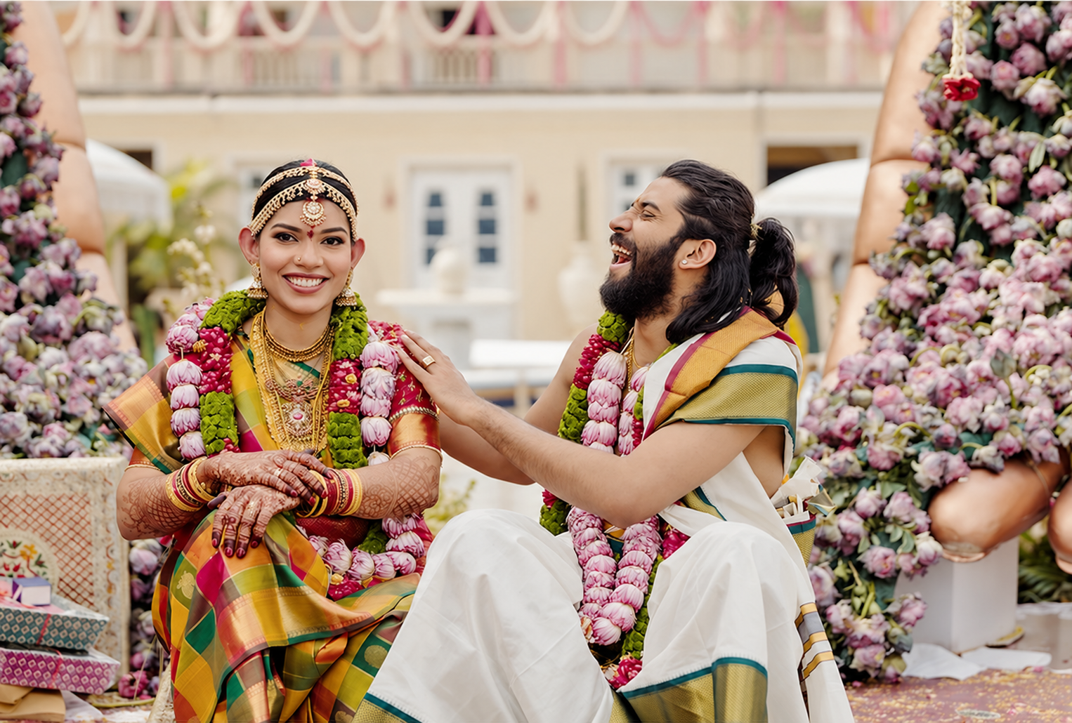 A bride in a traditional multicolored South Indian silk sari smiles forward while the groom in a white veshti laughs heartily during an outdoor wedding ceremony at Chomu Palace.