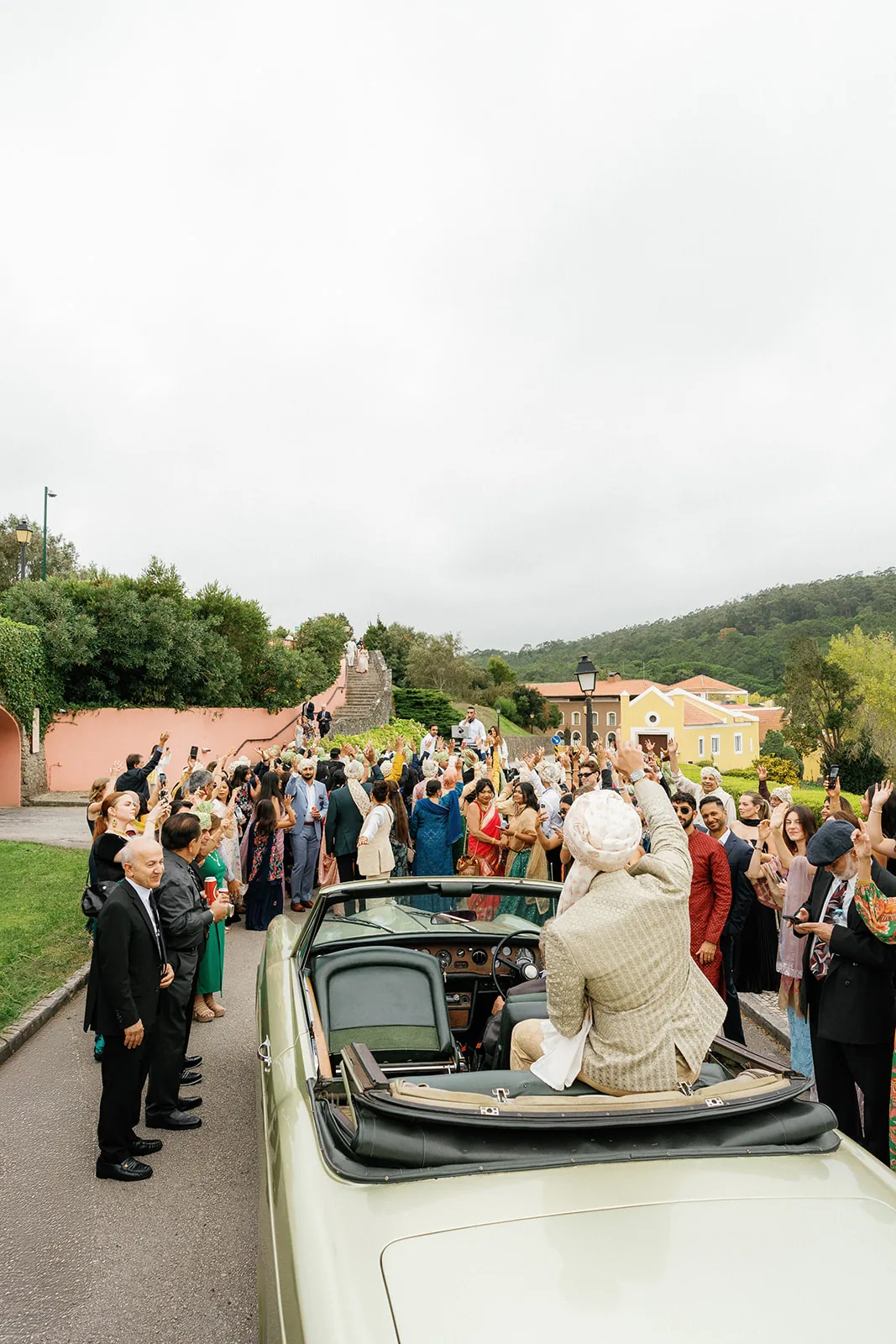 Groom arriving in a vintage convertible surrounded by dancing family and friends during an Indian baraat at Penha Longa Resort, Portugal.