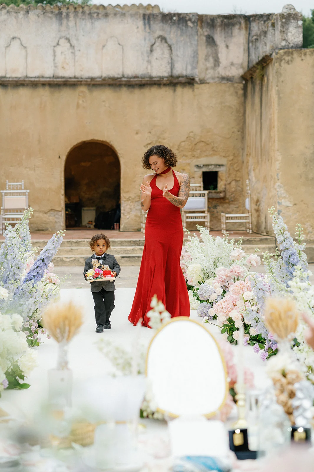 A young ring bearer carrying a decorated tray beside a guest in red on the pastel floral aisle at Penha Longa Resort, Portugal