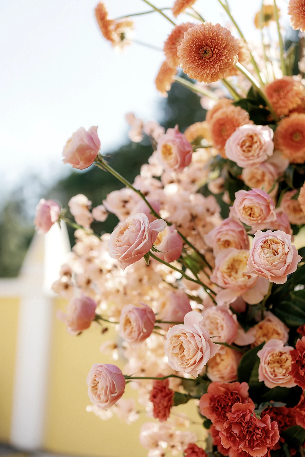 Peach and blush-pink roses with orange blooms, coral carnations and green foliage in a sunlit wedding floral arrangement.